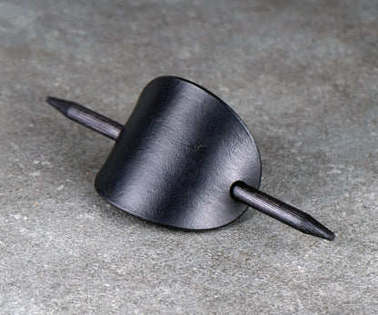 Black oval leather barrette with a stick threaded through both sides, resting on a gray textured surface.