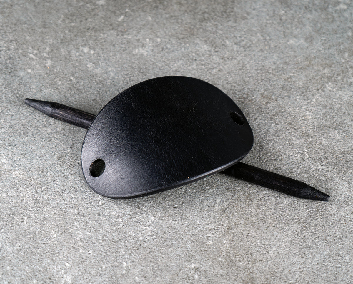 Black oval leather barrette with a stick threaded through, resting on a gray stone surface at a slight angle