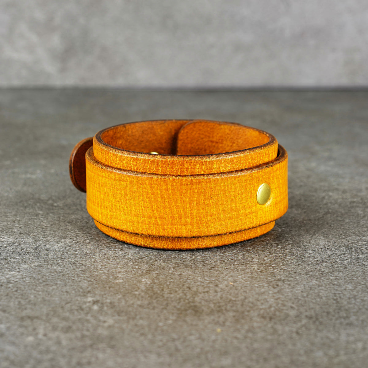 Yellow leather cuff bracelet standing upright on a gray surface, with a brass rivet visible on the front.