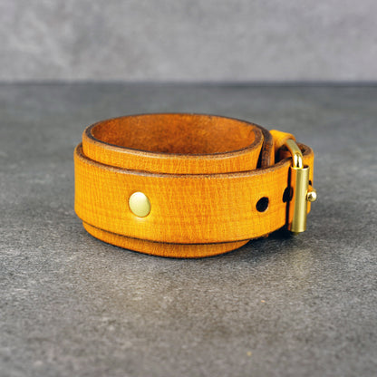 Yellow leather cuff bracelet coiled on a gray surface, showing a brass buckle, rivet, and punched holes.