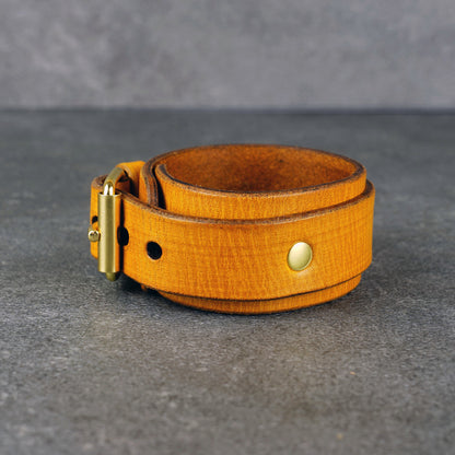 Yellow leather cuff bracelet coiled on a gray surface, showing a brass buckle, round rivet, and punched holes.