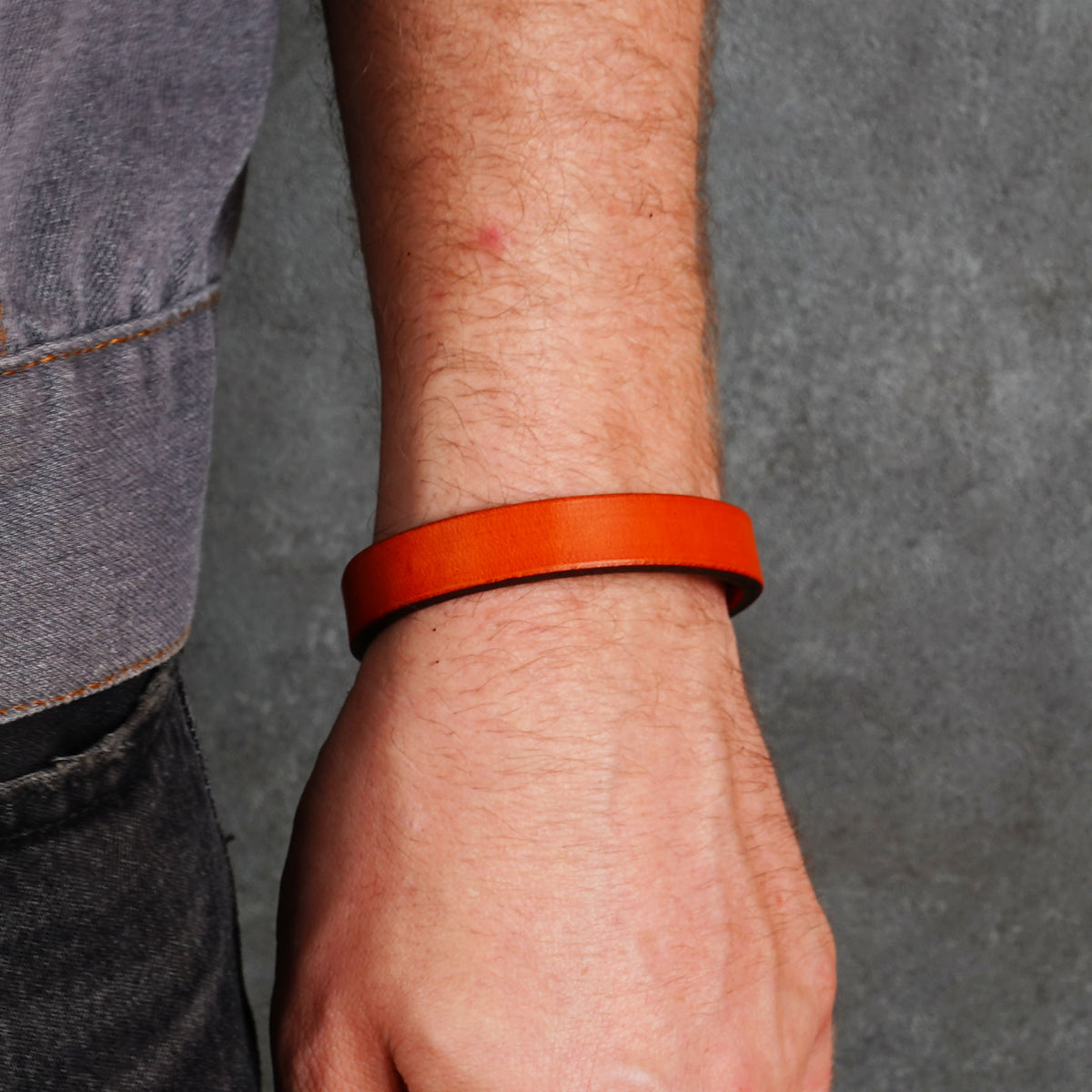 Bright orange leather bracelet worn on a wrist, shown in profile against a gray background.