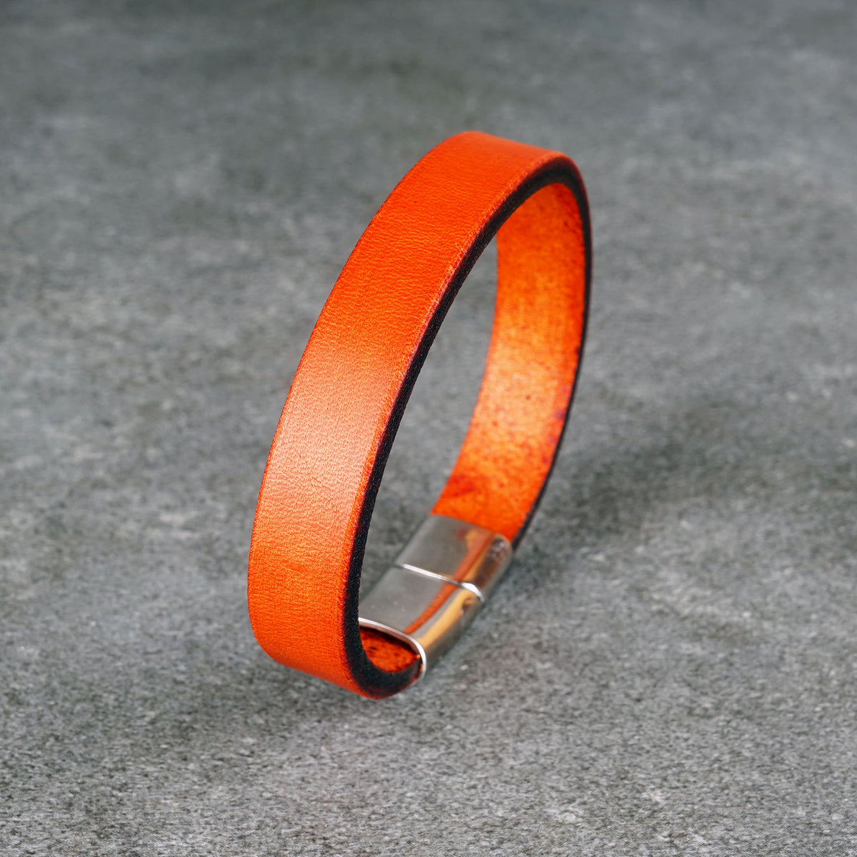 Bright orange leather bracelet standing upright in a loop on a gray surface, with a silver metal clasp at the base.