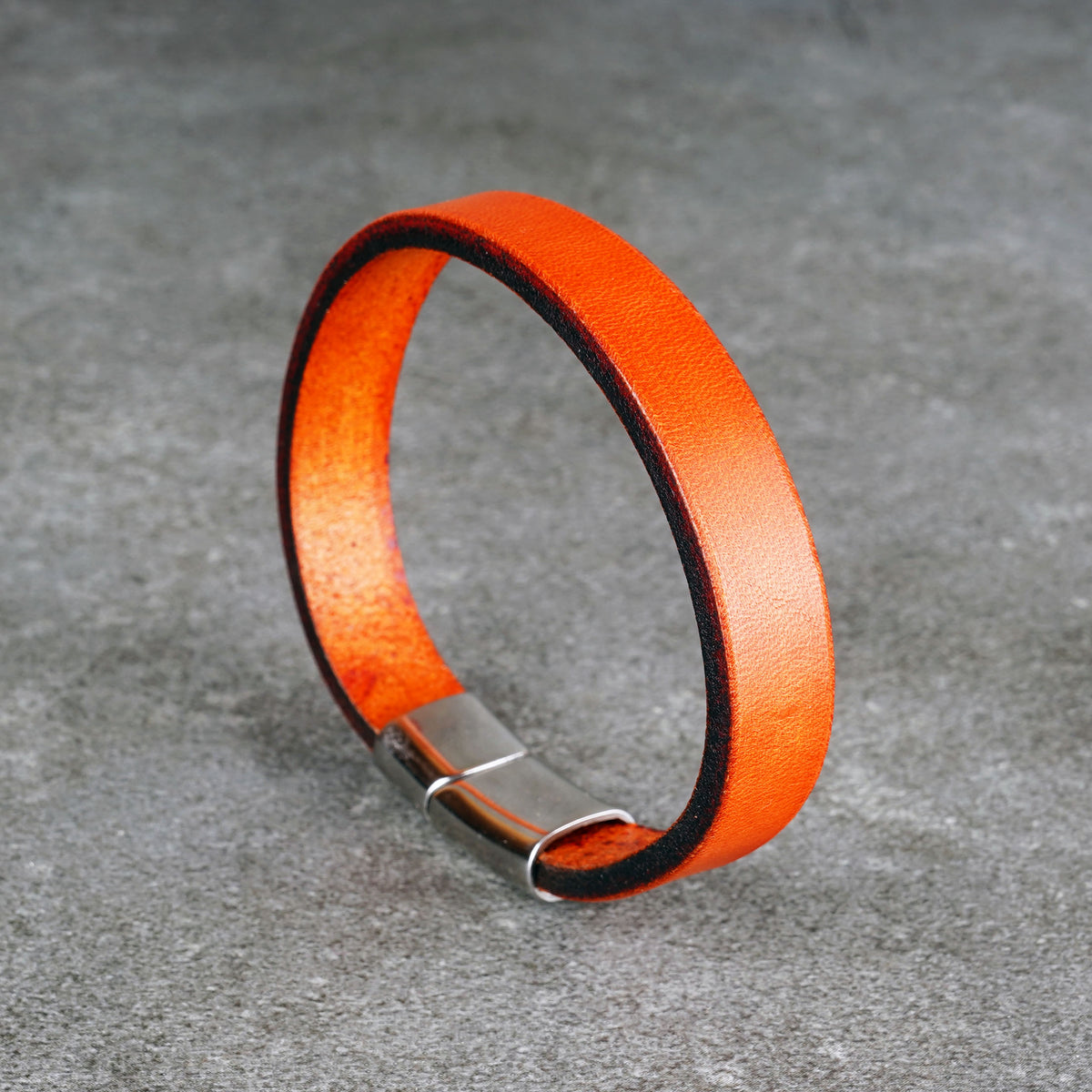 Orange leather bracelet standing upright in a loop on a gray surface, with a silver metal clasp at the bottom.