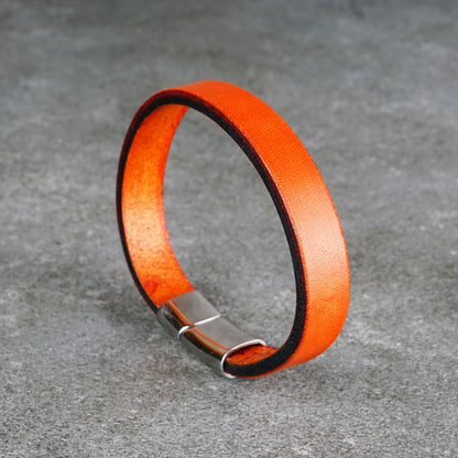Orange leather bracelet standing upright in a loop on a gray surface, with a silver metal clasp at the bottom.