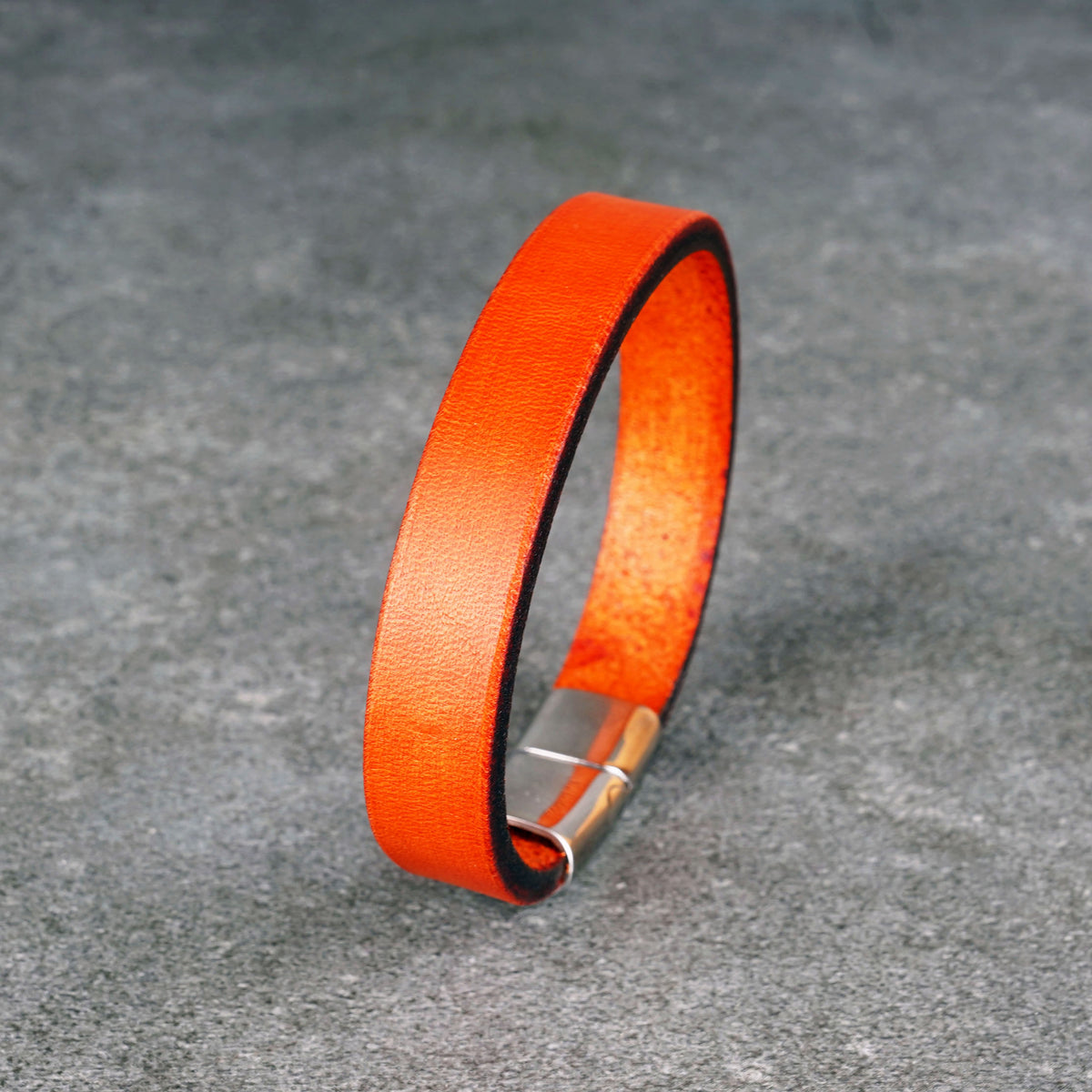 Orange leather bracelet standing upright in a loop on a gray surface, with a silver clasp at the bottom.
