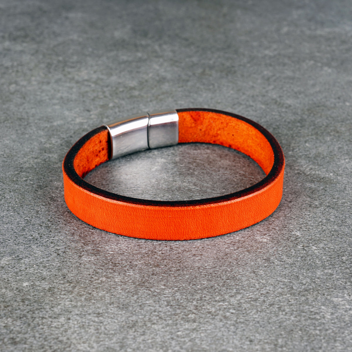 Orange leather bracelet laid in a circle on a gray surface, with a silver metal clasp at the top.