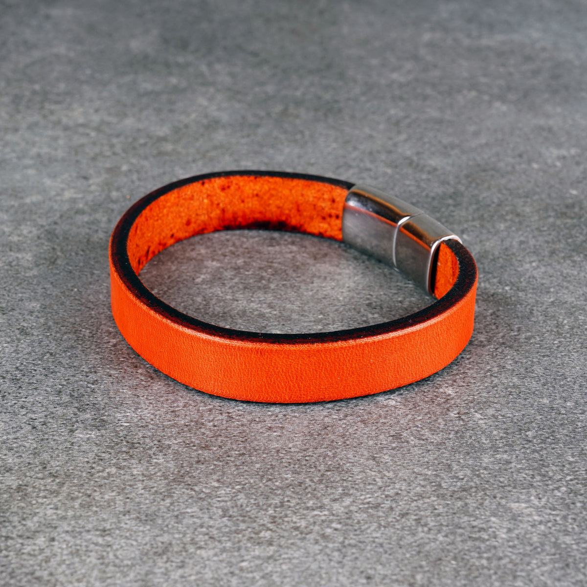 Orange leather bracelet laid in a circle on a gray surface, with a silver metal clasp at one side.