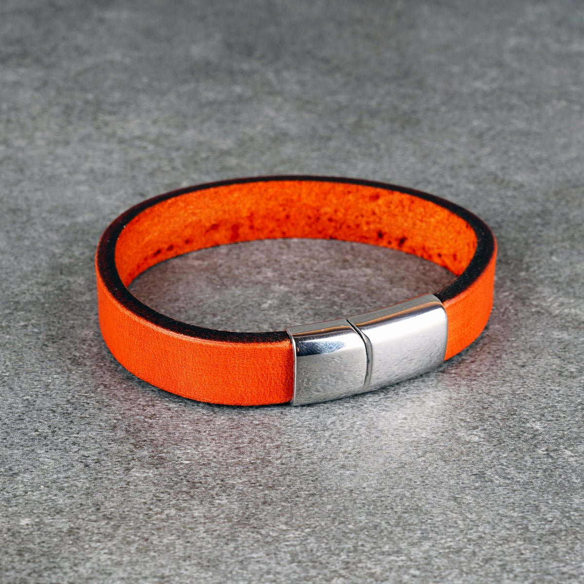 Orange leather bracelet with a silver-tone metal clasp, laid in a loop on a gray surface.