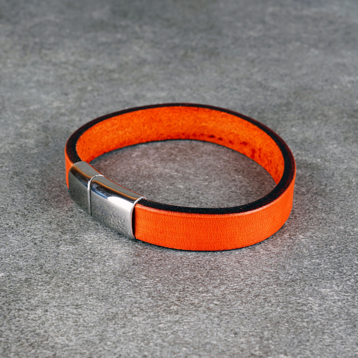 Orange leather bracelet laid in a loop on a gray surface, with a silver metal clasp at the front left.
