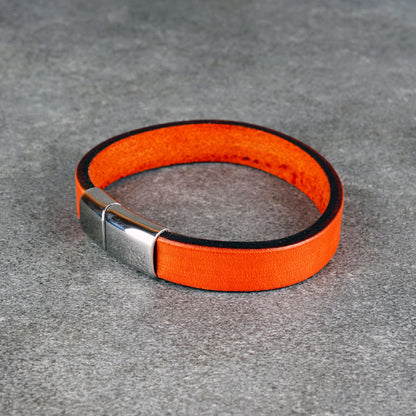Orange leather bracelet laid in a loop on a gray surface, with a silver metal clasp at the front left.