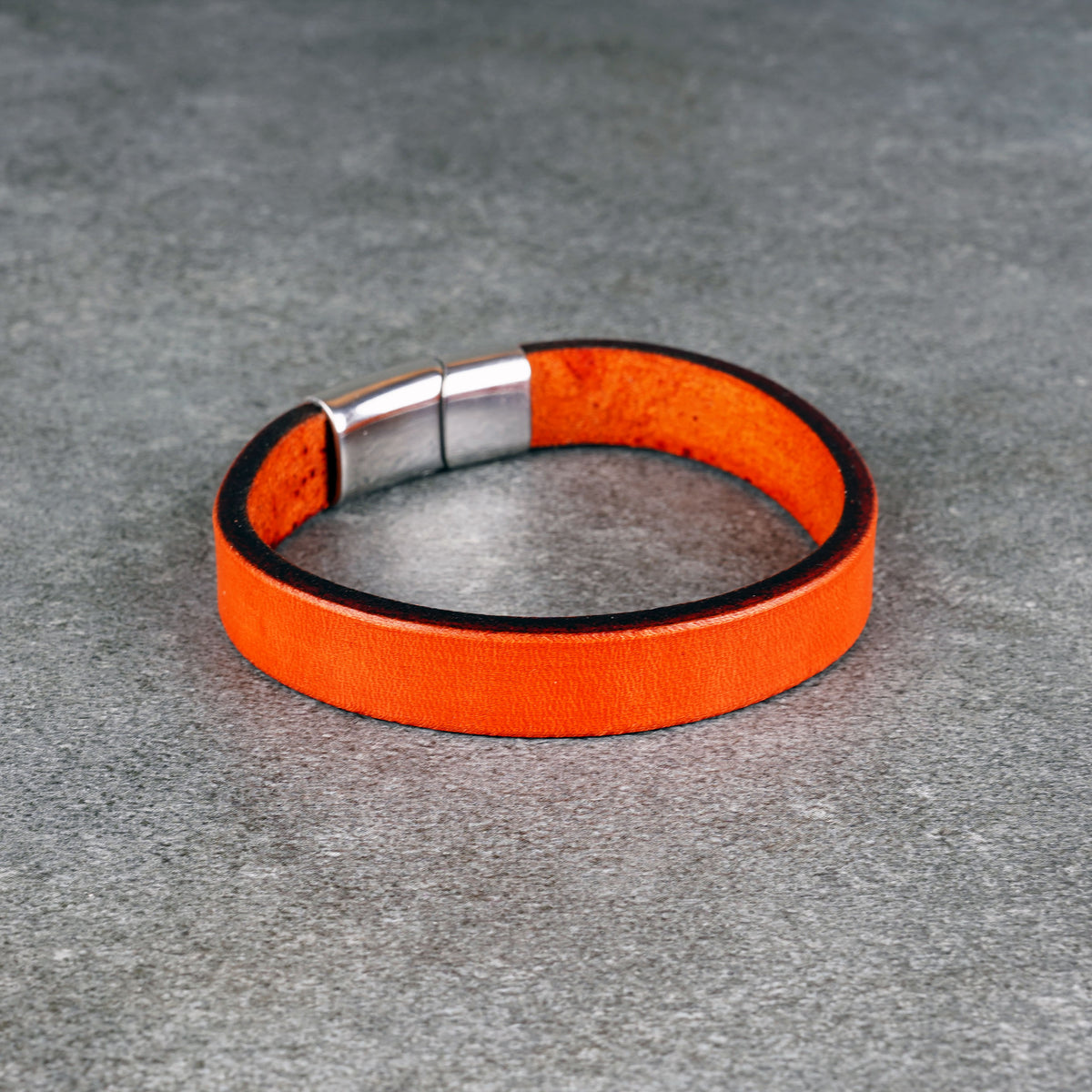Orange leather bracelet laid in a loop on a gray surface, with a silver metal clasp at the top left.